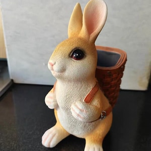May include: A ceramic bunny planter with a woven basket on its back. The bunny is light brown with pink-lined ears and paws. The basket is brown with a dark interior. The bunny has dark red eyes and is wearing brown straps.