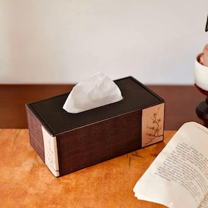 Puede incluir: Una caja de pañuelos rectangular marrón oscuro con una tapa negra. La caja tiene un acabado texturizado y un diseño floral en los laterales. Un pañuelo blanco sobresale de la parte superior. La caja está colocada sobre una superficie estampada.