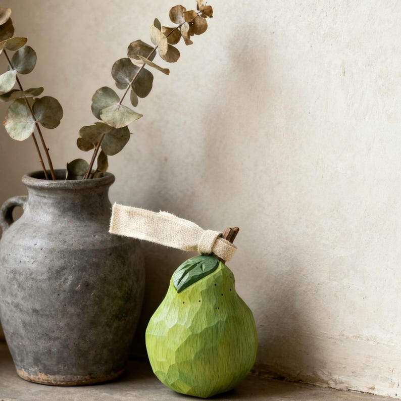 Peut inclure: Une poire en bois verte avec une feuille et une &eacute;tiquette en tissu beige attach&eacute;e &agrave; la tige. La poire pr&eacute;sente des d&eacute;tails sculpt&eacute;s et se trouve &agrave; c&ocirc;t&eacute; d'un vase en c&eacute;ramique grise avec des branches d'eucalyptus s&eacute;ch&eacute;es. Le fond est blanc cass&eacute; neutre.
