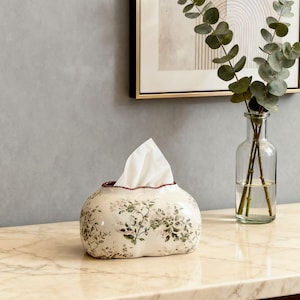 Op de afbeelding: Decoratieve keramische tissuebox met een bloemenpatroon in groen en roze. De off-white box heeft een geschulpte rand en een rode binnenkant. Witte tissues zijn zichtbaar. Een heldere glazen vaas met groen is op de achtergrond.
