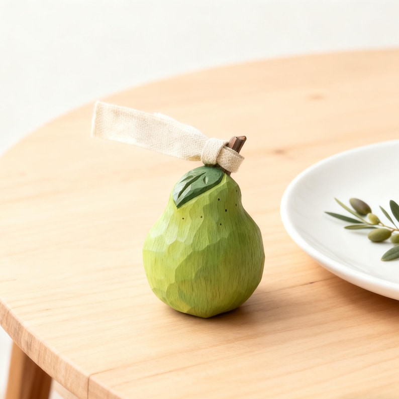 Peut inclure: Une figurine de poire en bois sculpt&eacute; &agrave; la main, dans des tons de vert, avec une petite feuille et un ruban beige nou&eacute; autour de la tige. La poire est pos&eacute;e sur une surface en bois clair, &agrave; c&ocirc;t&eacute; d'une assiette blanche avec des branches d'olivier.