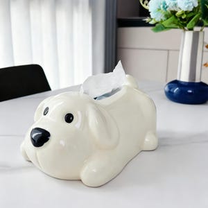 Op de afbeelding: Crèmekleurige keramische tissuebox in de vorm van een hond. De hond heeft zwarte ogen en een zwarte neus. Witte tissues komen uit de bovenkant van de rug van de hond. De tissuebox staat op een wit oppervlak.