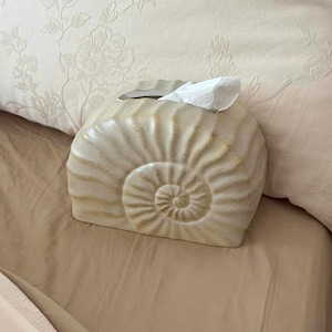 May include: A beige ceramic tissue box shaped like a nautilus shell, with a spiral design. The tissue box is on a bed with a beige comforter and a floral patterned pillow.