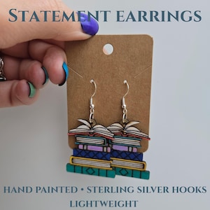 May include: Statement earrings depicting stacks of books in various colours, with an open book at the top. The earrings are hand-painted and have sterling silver hooks. The image text reads "STATEMENT EARRINGS", "HAND PAINTED • STERLING SILVER HOOKS", and "LIGHTWEIGHT".