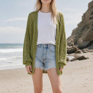 May include: An olive green open-front cardigan featuring a loose, airy knit pattern with long sleeves and ribbed cuffs, worn over a plain white t-shirt and light blue denim shorts with a frayed hemline, creating a layered casual outfit.