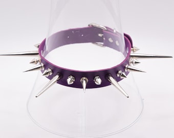 May include: A purple leather collar with silver metal spikes. The collar has a buckle closure and is displayed on a clear stand. The spikes are of varying sizes and shapes, adding a bold, edgy aesthetic.