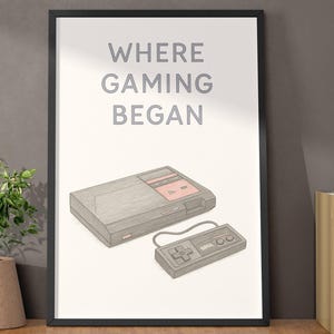 May include: Framed print featuring a retro video game console and controller. The print has the text "WHERE GAMING BEGAN" in gray. The console is gray with red accents. The print is displayed on a wooden shelf with a decorative vase, plant, and books.