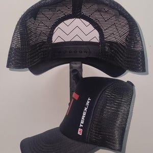 May include: Black trucker hat with a curved brim and mesh back. The hat features the text "TEREX.IRT" in white and red on the front. The back of the hat has a black adjustable snap closure.