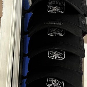 May include: Black trucker hats with a white logo that reads "THE BUNKER BUSTERS" above a shield design and "EST 2021". The hats are stacked, showing the front of the caps. The mesh back is visible.