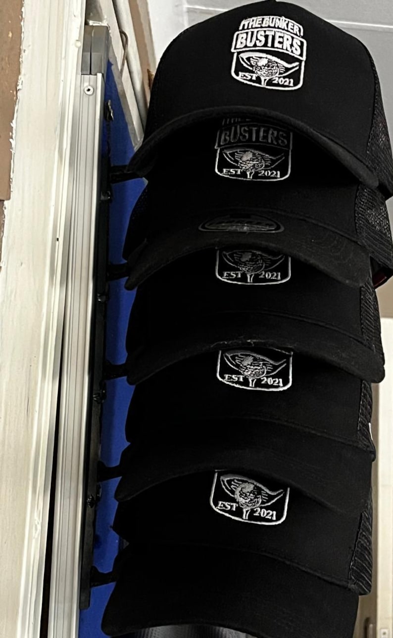 May include: Black trucker hats with a white logo that reads "THE BUNKER BUSTERS" above a shield design and "EST 2021". The hats are stacked, showing the front of the caps. The mesh back is visible.
