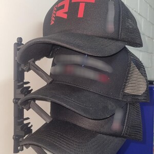 May include: A stack of black trucker hats with a red logo on the front. The hats have a mesh back and a curved brim. The hats are displayed on a black hat rack against a white wall.
