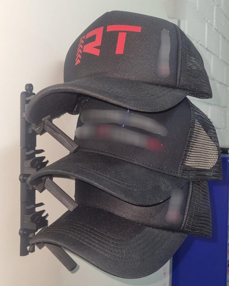 May include: A stack of black trucker hats with a red logo on the front. The hats have a mesh back and a curved brim. The hats are displayed on a black hat rack against a white wall.