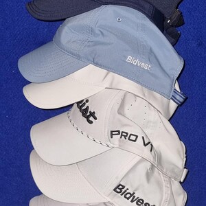 May include: A stack of baseball caps in various colors, including navy blue, light blue, and white. Some caps have the "Bidvest" or "Titleist" logo embroidered on the front. The caps are made of a textured material.