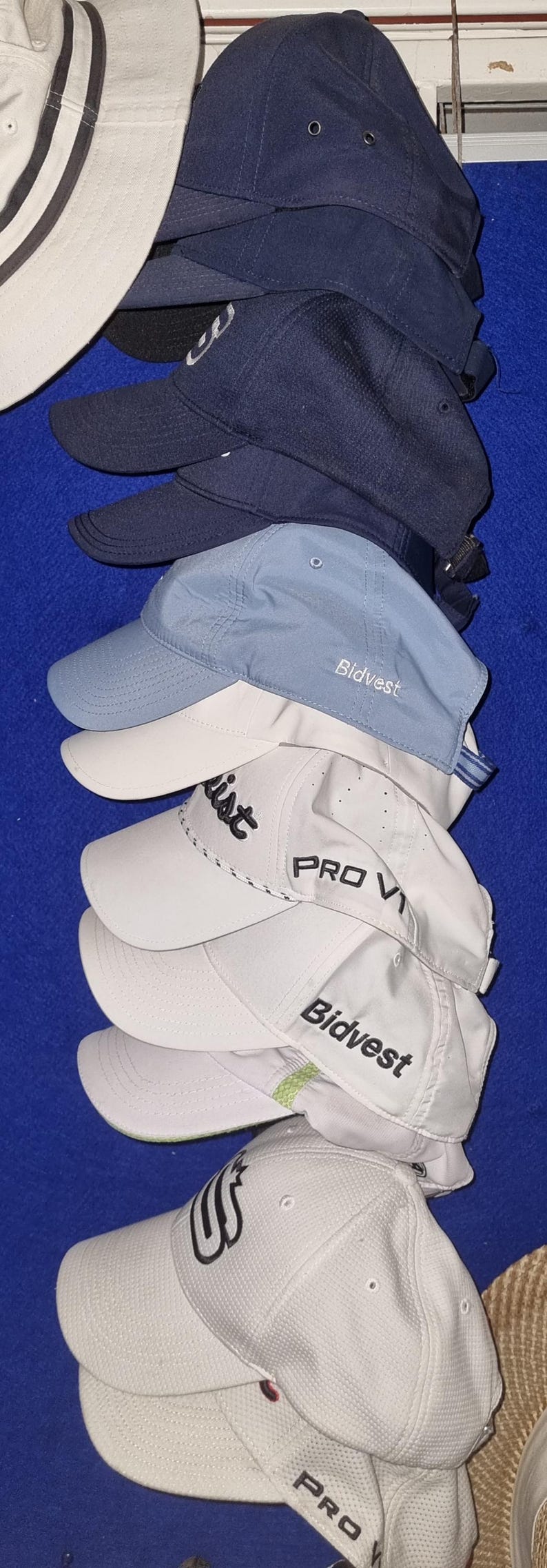 May include: A stack of baseball caps in various colors, including navy blue, light blue, and white. Some caps have the "Bidvest" or "Titleist" logo embroidered on the front. The caps are made of a textured material.
