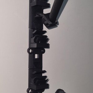 May include: Black 3D-printed wall-mounted rack with multiple angled holders and cylindrical supports. The rack has a series of geometric shapes and a loop at the top for hanging. It is designed to hold items.