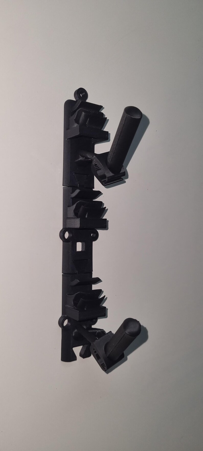 May include: Black 3D-printed wall-mounted rack with multiple angled holders and cylindrical supports. The rack has a series of geometric shapes and a loop at the top for hanging. It is designed to hold items.