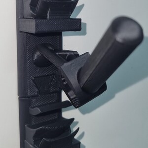 May include: A black 3D-printed wall-mounted holder with multiple slots and a protruding handle. The holder is designed to organize and store items, featuring a textured surface and a loop for hanging. The design is modern and functional.