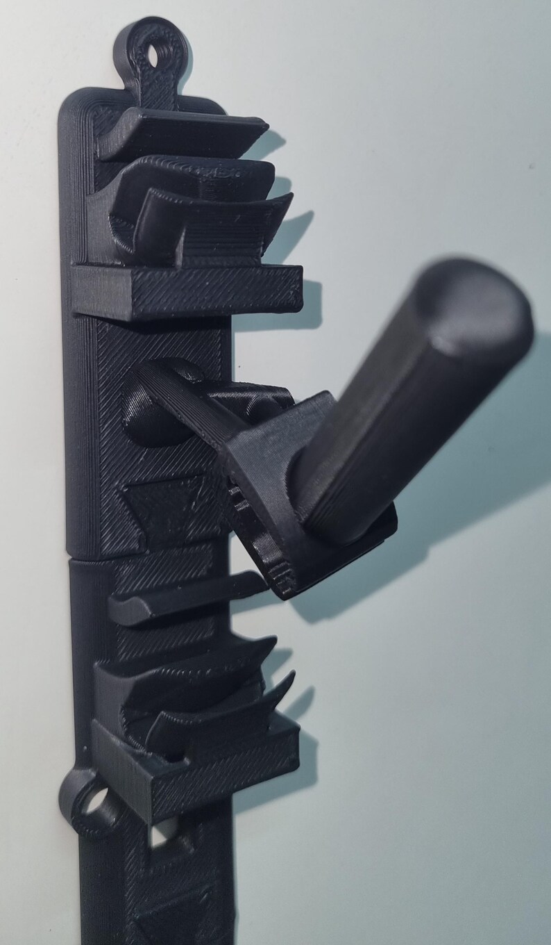 May include: A black 3D-printed wall-mounted holder with multiple slots and a protruding handle. The holder is designed to organize and store items, featuring a textured surface and a loop for hanging. The design is modern and functional.