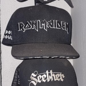 May include: A collection of black baseball caps with various band logos embroidered in white. The caps include logos for "The Doors", "The Cure", "Iron Maiden", "Seether", and "Rammstein".