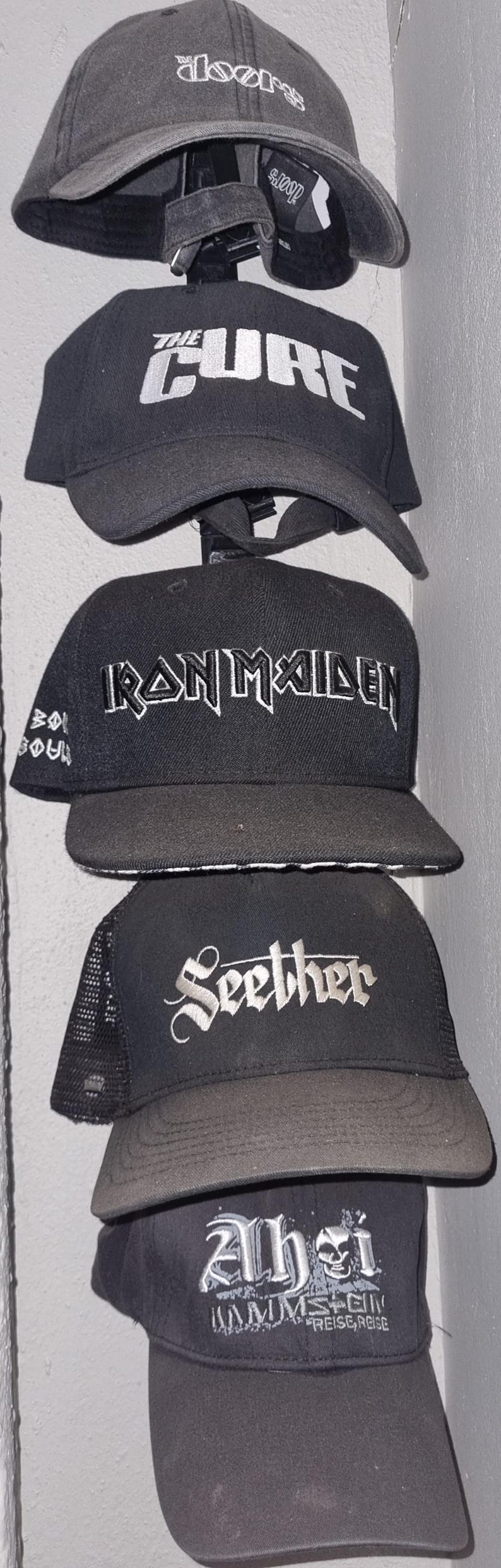 May include: A collection of black baseball caps with various band logos embroidered in white. The caps include logos for "The Doors", "The Cure", "Iron Maiden", "Seether", and "Rammstein".
