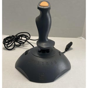 Puede incluir: Un joystick Logitech Wingman gris oscuro con un botón naranja. El joystick tiene un mango curvo y una base ancha. Un cable y un enchufe negros se extienden desde la base.