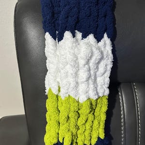 May include: A chunky knit blanket in a color block design. The top and bottom sections are navy blue, with a white center stripe and a bright green stripe. The blanket is made of a soft, textured yarn.
