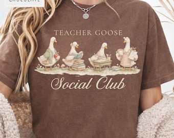 May include: A chocolate brown t-shirt featuring a graphic of four geese engaged in school activities. The text reads "TEACHER GOOSE" above the geese and "Social Club" below. The geese are depicted with books, a desk, and a coffee mug.