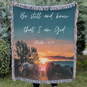 May include: A woven throw blanket featuring a sunset scene with horses grazing in a field. The blanket has a teal background with the text "Be still and know that I am God" and "Psalm 46:10" in white script. The edges are fringed.
