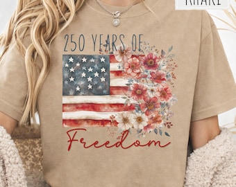 May include: A khaki t-shirt featuring a graphic print of the American flag combined with a floral arrangement. The text reads 250 YEARS OF at the top and Freedom in red script at the bottom. The design includes red, white, and blue colors.