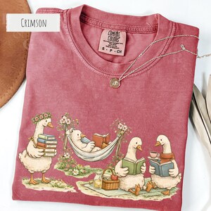 May include: Crimson-colored t-shirt featuring a whimsical illustration of geese reading books and relaxing. One goose is in a hammock, while others are enjoying tea and books. A silver necklace with a pendant is also visible.