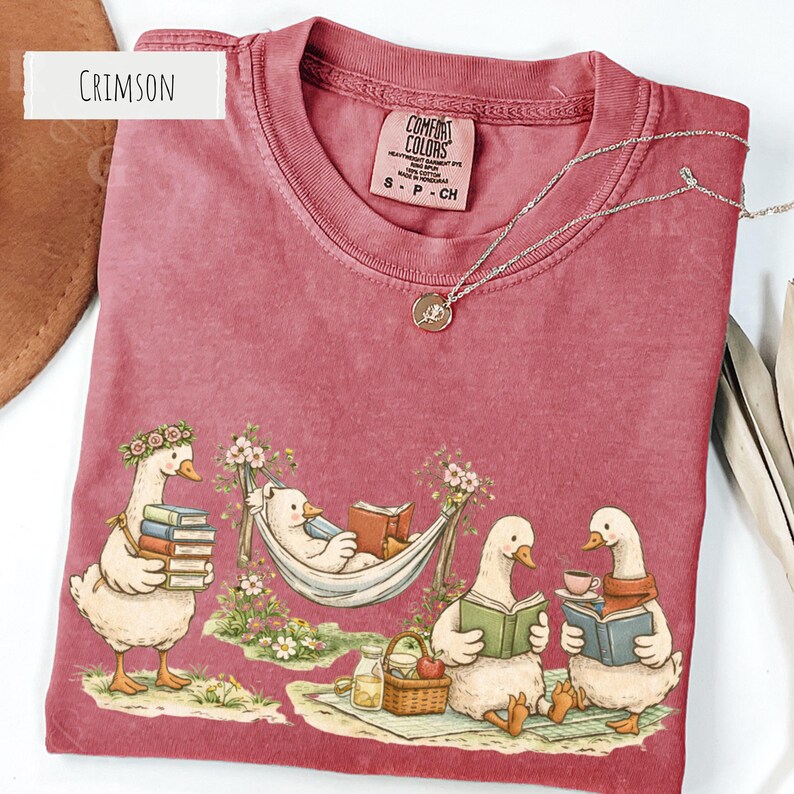 May include: Crimson-colored t-shirt featuring a whimsical illustration of geese reading books and relaxing. One goose is in a hammock, while others are enjoying tea and books. A silver necklace with a pendant is also visible.