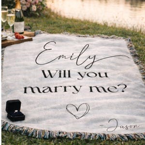 May include: A light-colored woven throw blanket with fringed edges featuring the black printed text "Emily Will you marry me?" followed by a hand-drawn heart and "- Jason". A small black ring box containing a ring sits on the blanket.