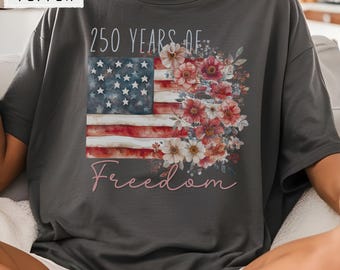 May include: A charcoal gray t-shirt featuring a graphic print of the American flag combined with a cluster of watercolor-style flowers. The text 250 YEARS OF appears above the flag, and the word Freedom is printed in a script font below it.