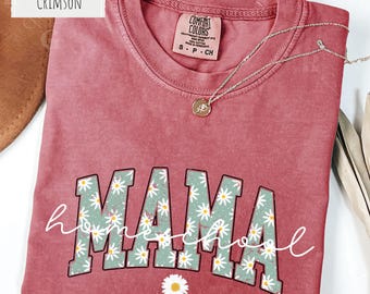 May include: A crimson short-sleeve t-shirt with a crew neckline. The front features the word MAMA in large block letters filled with a green and white daisy floral pattern, overlaid with the word homeschool in white cursive script.