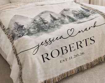 May include: A cream-colored woven throw blanket featuring a watercolor-style mountain and pine forest landscape. The text reads jessica & mark, ROBERTS, and EST 11.20.26. The blanket has a decorative fringed edge along the bottom and sides.
