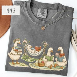 May include: A gray Comfort Colors t-shirt featuring a whimsical illustration of geese reading books and enjoying a picnic. The shirt has a crew neck and short sleeves. A silver necklace with a pendant is on the shirt. The word "Pepper" is on the top left.