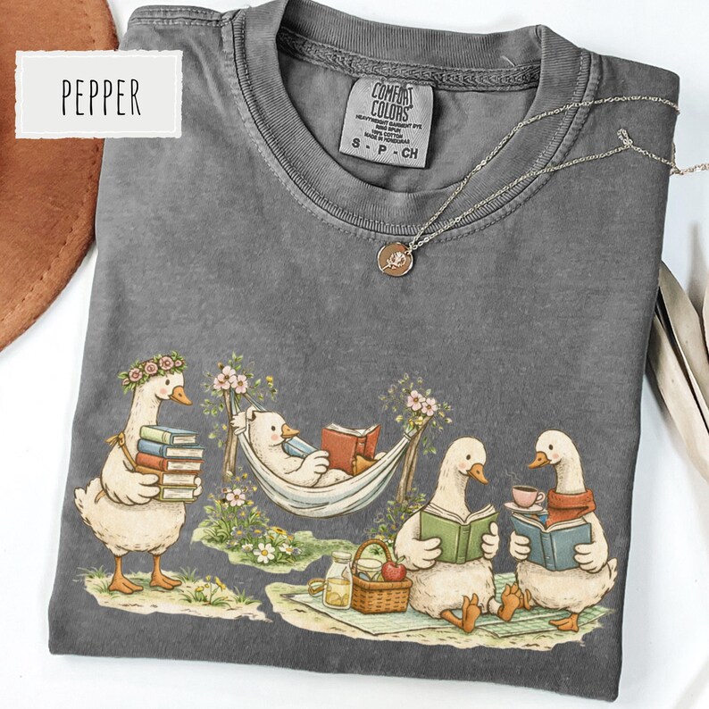 May include: A gray Comfort Colors t-shirt featuring a whimsical illustration of geese reading books and enjoying a picnic. The shirt has a crew neck and short sleeves. A silver necklace with a pendant is on the shirt. The word "Pepper" is on the top left.