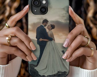 May include: A rectangular phone case featuring a printed photograph of a couple in formal wedding attire standing together outdoors at sunset. The case has a cutout for a dual-lens camera system and is held by hands with manicured nails.