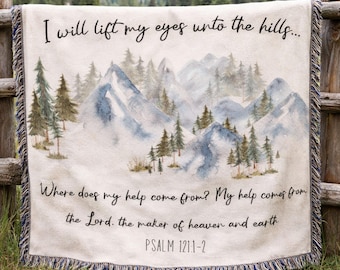 May include: A cream-colored woven throw blanket featuring a watercolor-style mountain and pine forest landscape. The text reads: I will lift my eyes unto the hills... Where does my help come from? My help comes from the Lord, the maker of heaven and earth. PSALM 121:1-2. The blanket has fringed edges.