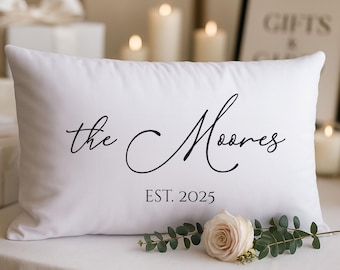 May include: A rectangular white throw pillow featuring black script text that reads the Moores and the text EST. 2025 printed below in a simple serif font. The pillow is positioned with a single pale pink rose and green leaves in front.