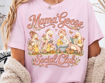 May include: A light pink t-shirt featuring a graphic illustration of geese and ducklings with the text "Mama Goose Social Club". The design includes geese engaged in activities like drinking from a mug, bathing, and reading a book, surrounded by flowers.