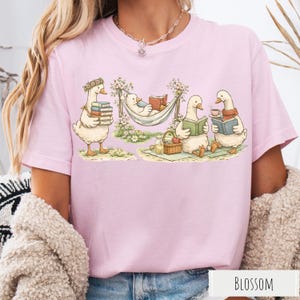 May include: Light pink t-shirt featuring a whimsical illustration of geese reading books. One goose carries books, another relaxes in a hammock, and two more enjoy reading and tea. The word "BLOSSOM" is printed at the bottom.