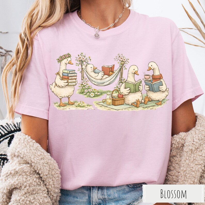 May include: Light pink t-shirt featuring a whimsical illustration of geese reading books. One goose carries books, another relaxes in a hammock, and two more enjoy reading and tea. The word "BLOSSOM" is printed at the bottom.