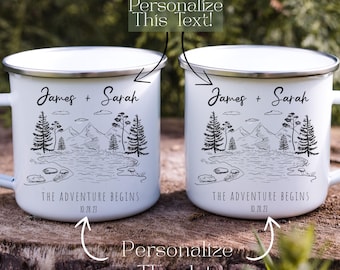 May include: Two white enamel mugs with silver rims, featuring a black and white mountain and lake landscape design. The text "James + Sarah" and "THE ADVENTURE BEGINS 10.28.23" are printed on the mugs. The mugs are sitting on a wooden surface.