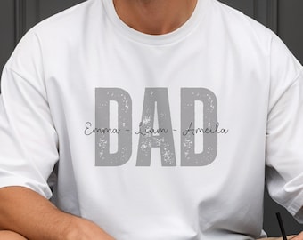 May include: A white short-sleeved t-shirt featuring the word DAD printed in large, distressed grey block letters. The names Emma, Liam, and Amelia are written in a thin, black cursive script across the center of the grey letters.