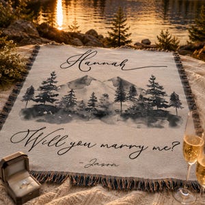 May include: A woven throw blanket featuring a watercolor-style mountain and pine tree landscape. The text reads Hannah at the top and Will you marry me? - Jason at the bottom. A ring box with a ring and a champagne flute are placed nearby.