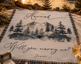 May include: A woven throw blanket featuring a watercolor-style mountain and pine tree landscape. The text reads Hannah at the top and Will you marry me? - Jason at the bottom. A ring box with a ring and a champagne flute are placed nearby.