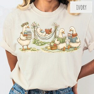 May include: Ivory-colored t-shirt featuring a whimsical illustration of geese reading books. One goose carries books, another relaxes in a hammock, and two others read with drinks. A picnic basket and flowers complete the scene.