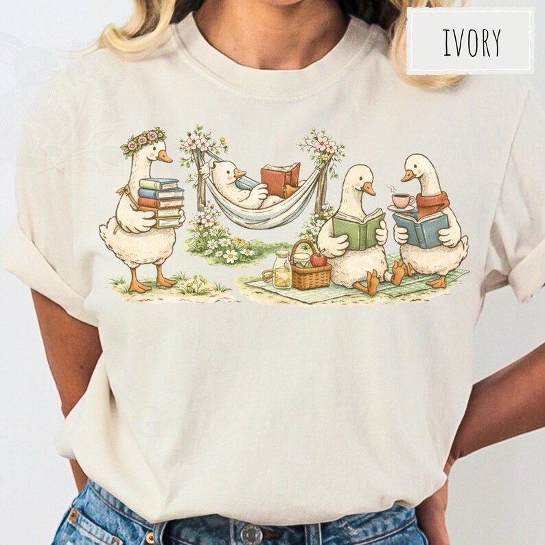 May include: Ivory-colored t-shirt featuring a whimsical illustration of geese reading books. One goose carries books, another relaxes in a hammock, and two others read with drinks. A picnic basket and flowers complete the scene.