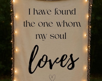 May include: A cream-colored woven throw blanket with fringed edges featuring the black printed text I have found the one whom my soul loves and a small hand-drawn heart outline centered at the bottom, illuminated by a string of warm fairy lights.